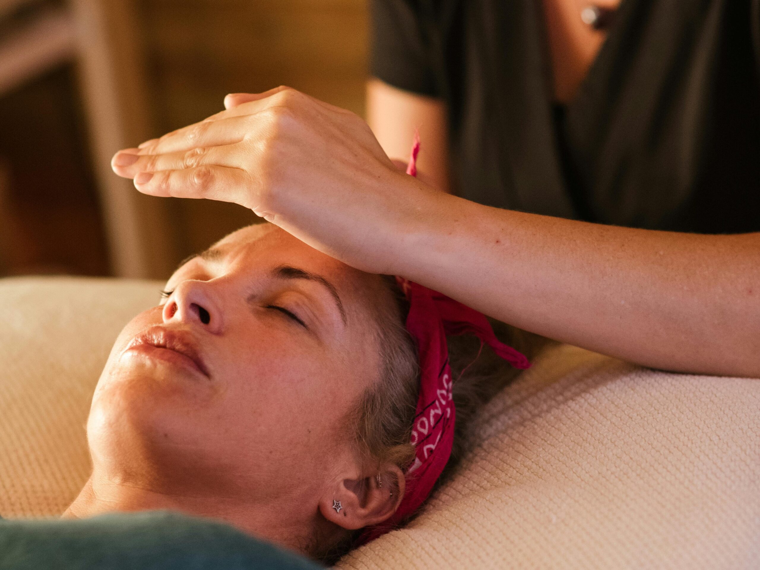 Relaxed woman with closed eyes lying under blanket while getting reiki therapy treatment from unrecognizable therapist in cozy salon with blurred background